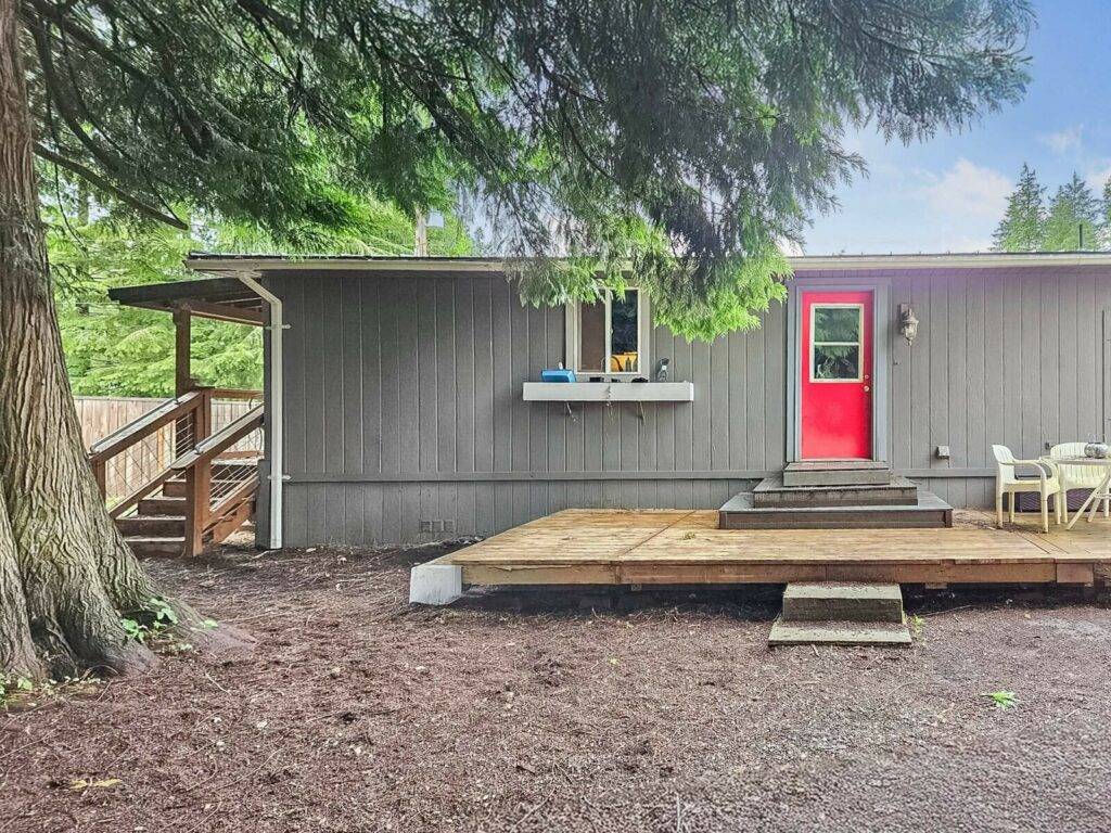 Back deck and red entry door of the Concrete, WA manufactured home, shaded by tall evergreens
